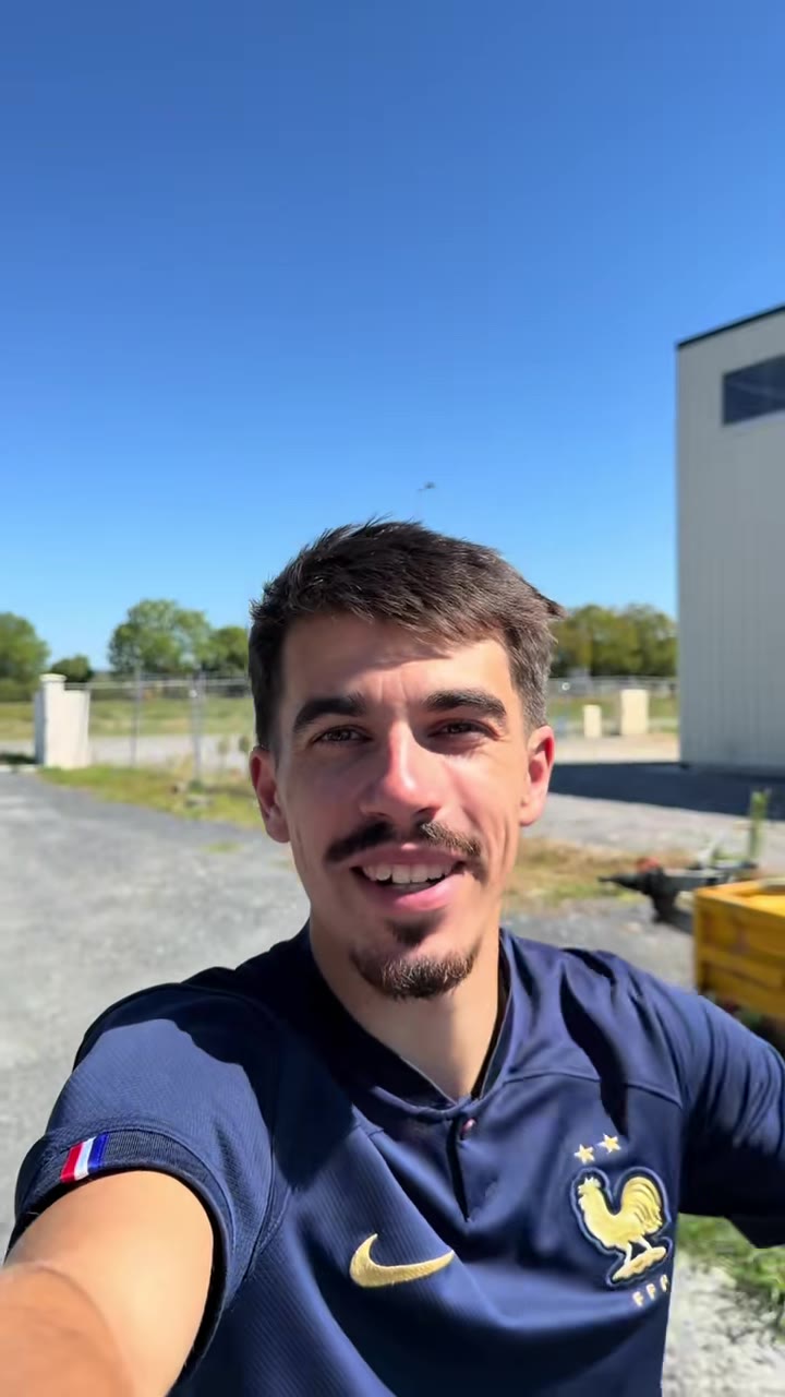 Alexis souriant devant un bâtiment et un chemin de gravier, lors du tournage de la vidéo à Lons