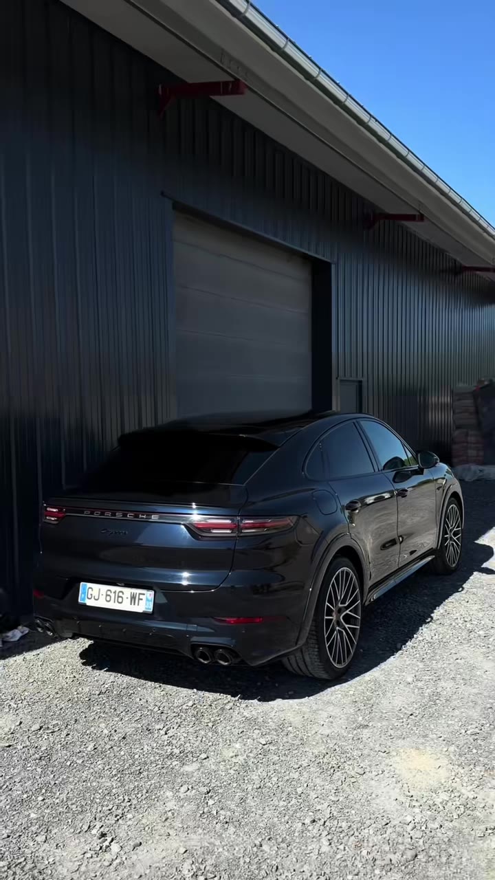 Porsche Cayenne noire vue de trois quarts arrière, garée sur du gravier devant un bâtiment sombre à Lons